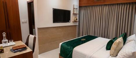 Quarto de hotel com cama, escrivaninha com cadeira, televisão na parede e janela com cortinas.