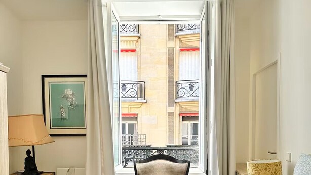 Ein Hotelzimmer mit Bett, Sessel, Schreibtisch, Lampe und Blick auf ein Gebäude mit Balkonen.