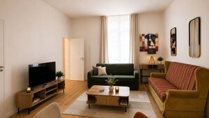 Ein modernes Wohnzimmer mit Fernseher, einem grünen Sofa, einem gestreiften Sofa, einem Holztisch und einem kleinen Beistelltisch mit einer Pflanze.