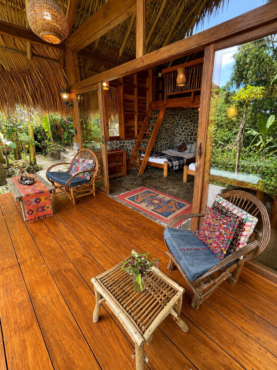 Une terrasse en bois avec des meubles en osier, un mur en pierre et un toit de chaume.