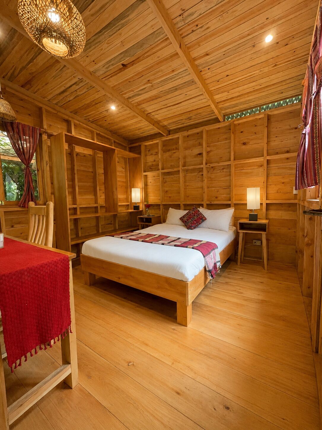 A wooden cabin-style bedroom with a large bed, two bedside tables, a chair, and a hanging light fixture.