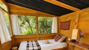 Un dormitorio acogedor de estilo cabaña con cama, una mesita de noche pequeña, una lámpara y vistas al follaje exterior.