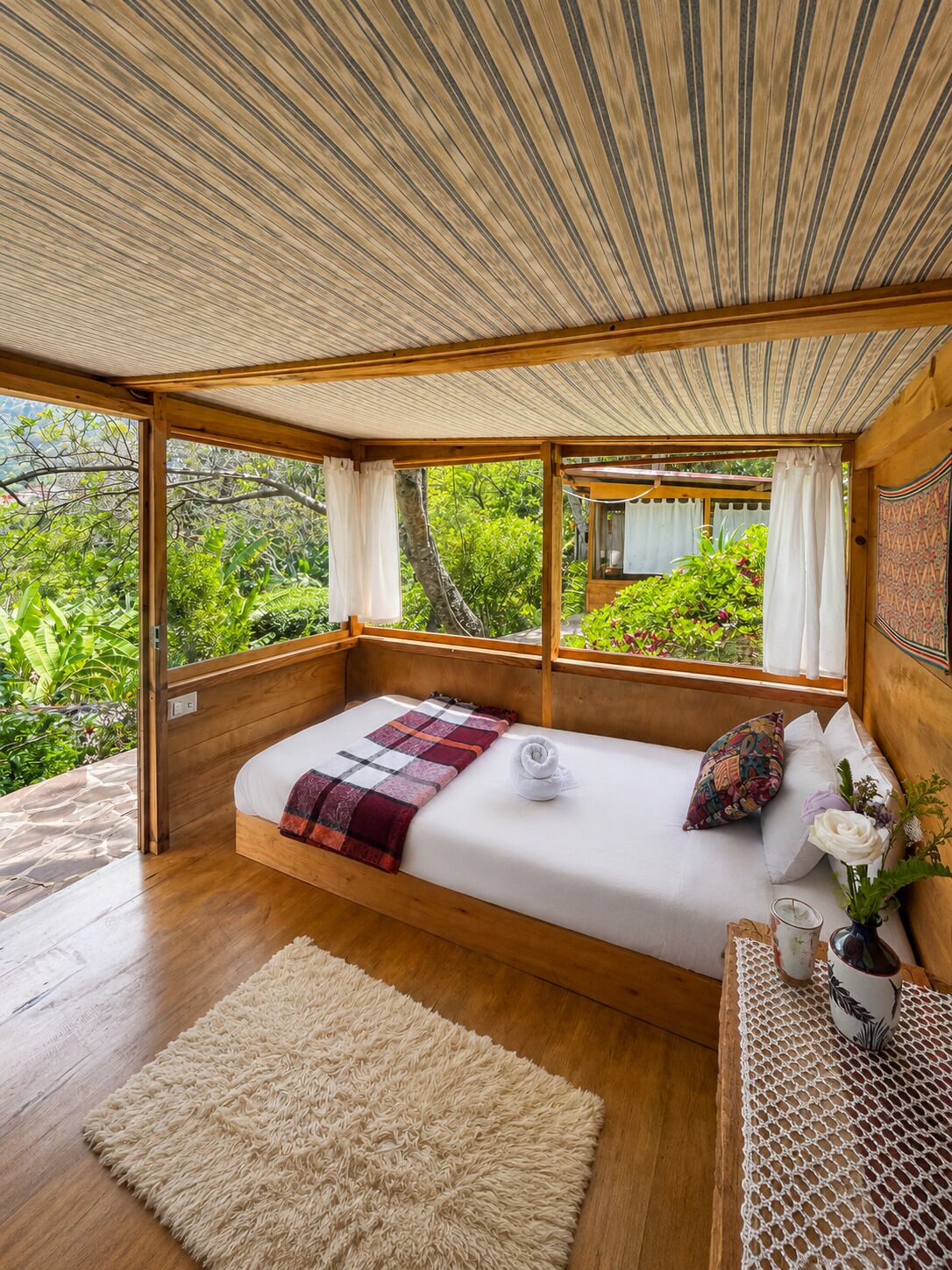 A bedroom with a wooden bed, a view of greenery, and a woven rug.
