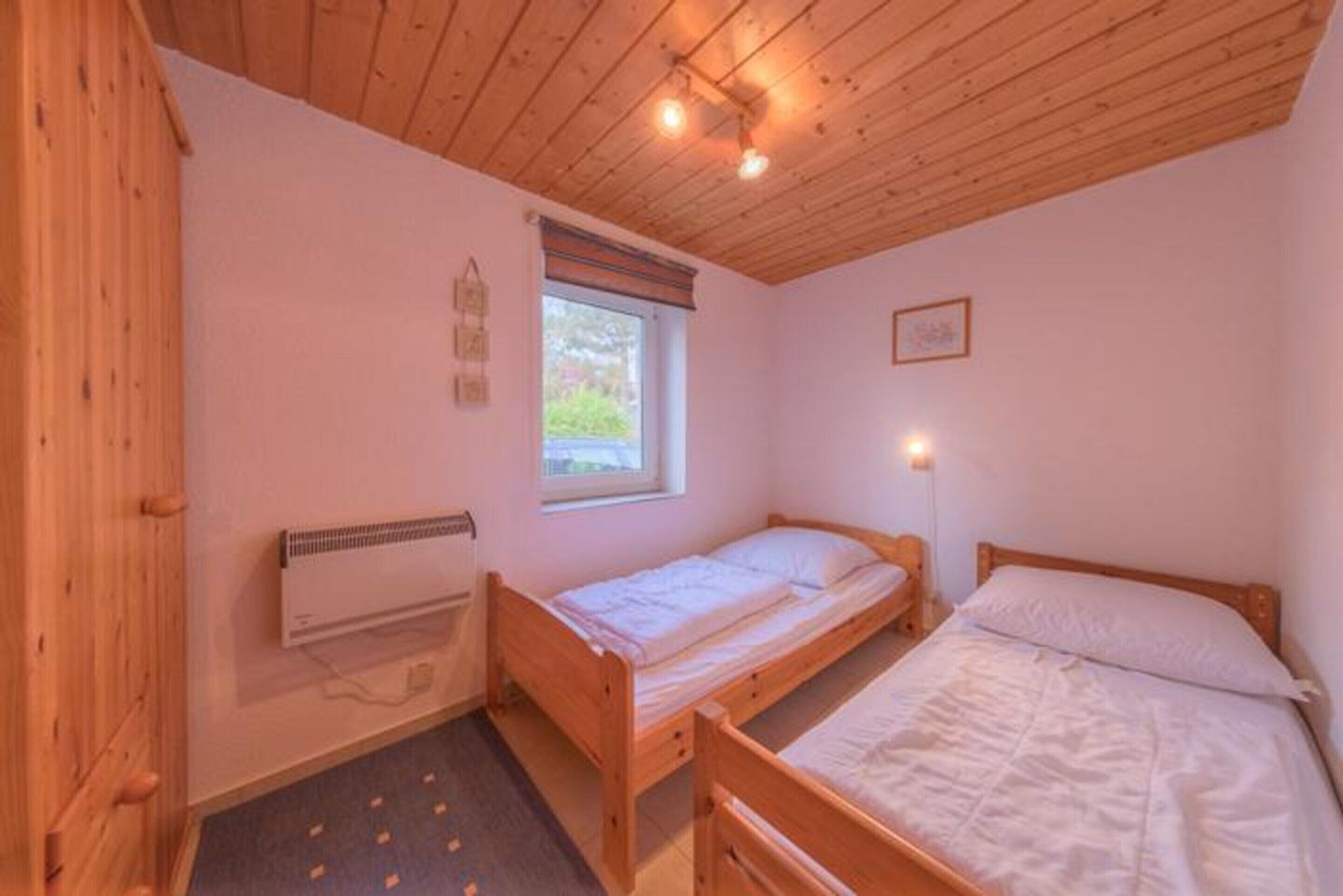 A small, single-room accommodation with a wooden ceiling, a window with a view, a bed, a bedside table, and a wall-mounted heater.
