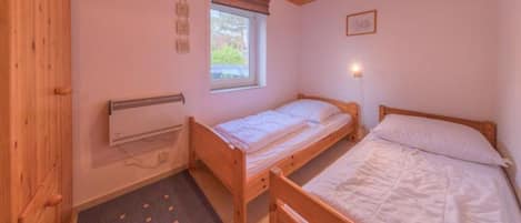 A small, single-room accommodation with a wooden ceiling, a window with a view, a bed, a bedside table, and a wall-mounted heater.