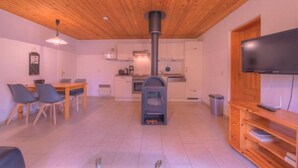 Un salon avec une cheminée, un coin repas avec des chaises, un téléviseur posé sur un meuble en bois, et une cuisine à l’arrière-plan.
