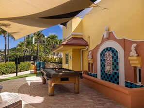 A pool table in an outdoor area with a yellow building, palm trees, and a covered seating area. - Marriott's Frenchman's Cove 2 Bedroom Villa (St Thomas)