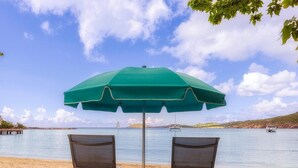 Twee strandstoelen onder een groene parasol op een zandstrand met een rustige zee en boten in de verte.