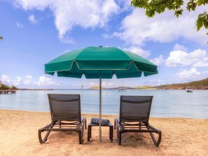 Two beach chairs under a green umbrella on a sandy beach with a calm sea and boats in the distance. - Marriott's Frenchman's Cove 2 Bedroom Villa (St Thomas)