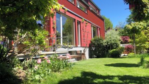 Ein rotes Gebäude mit großen Fenstern, einem Balkon und einem gepflegten Garten mit rosa Blüten.