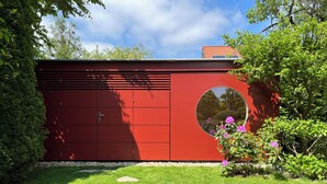 A red garage with a circular window, surrounded by green grass and bushes.