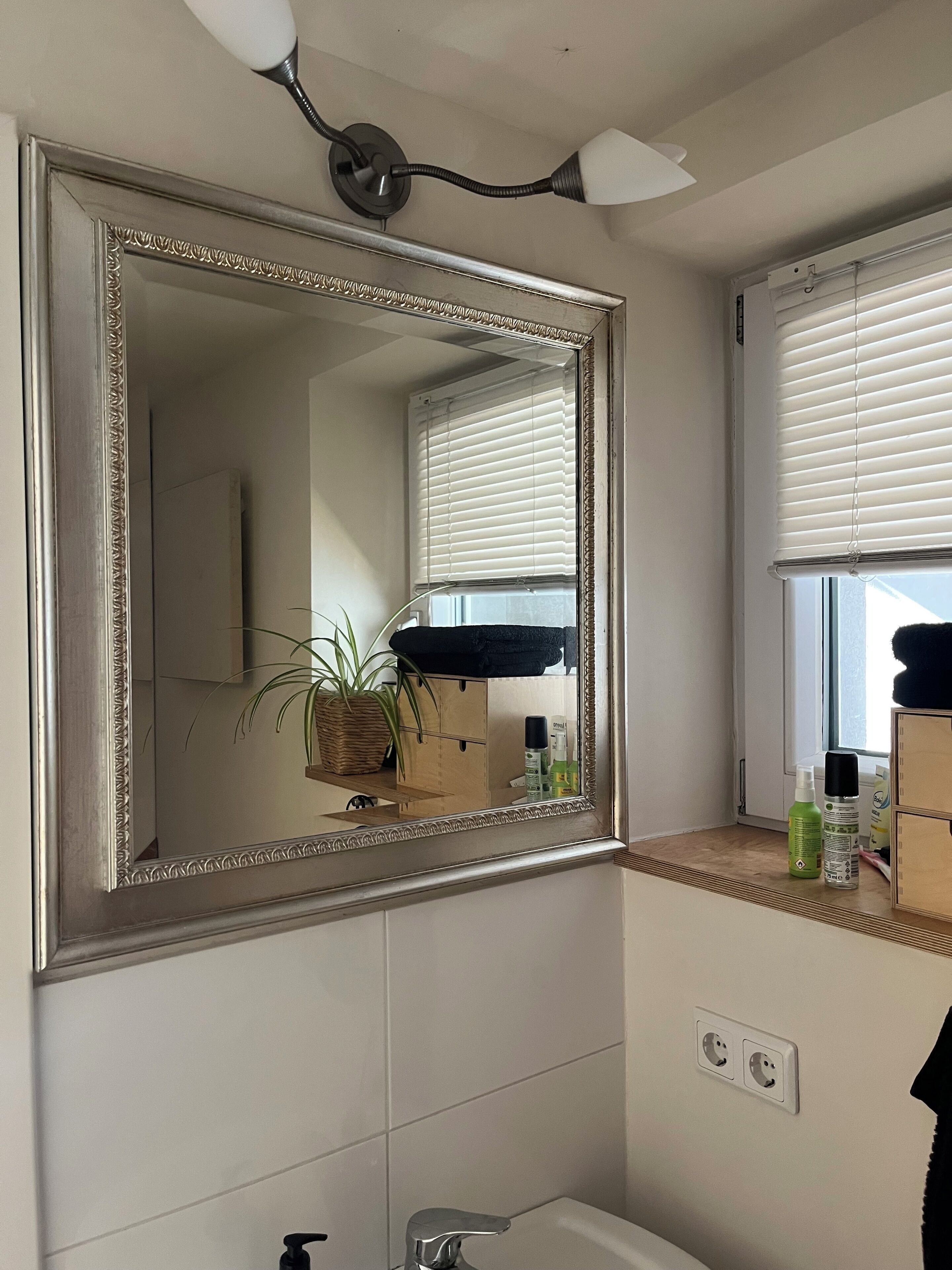 Une salle de bain dotée d’un grand miroir, d’un luminaire fixé au mur, d’une fenêtre avec des stores et d’un lavabo avec un robinet.