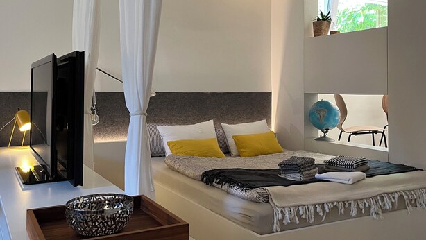 A modern bedroom with a bed, bedside table, lamp, and a window with a view of greenery.
