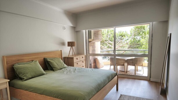 Een slaapkamer met een houten bed, een nachtkastje, een dressoir, een lamp en uitzicht op het landschap door een groot raam.