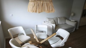 Een moderne woonkamer met een salontafel met glazen blad, omringd door witte stoelen, een grijze bank en een geweven lamp.