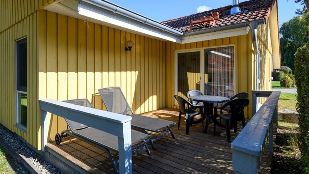 Um edifício amarelo com um alpendre coberto, com mesa e cadeiras, um pequeno deck e uma janela.