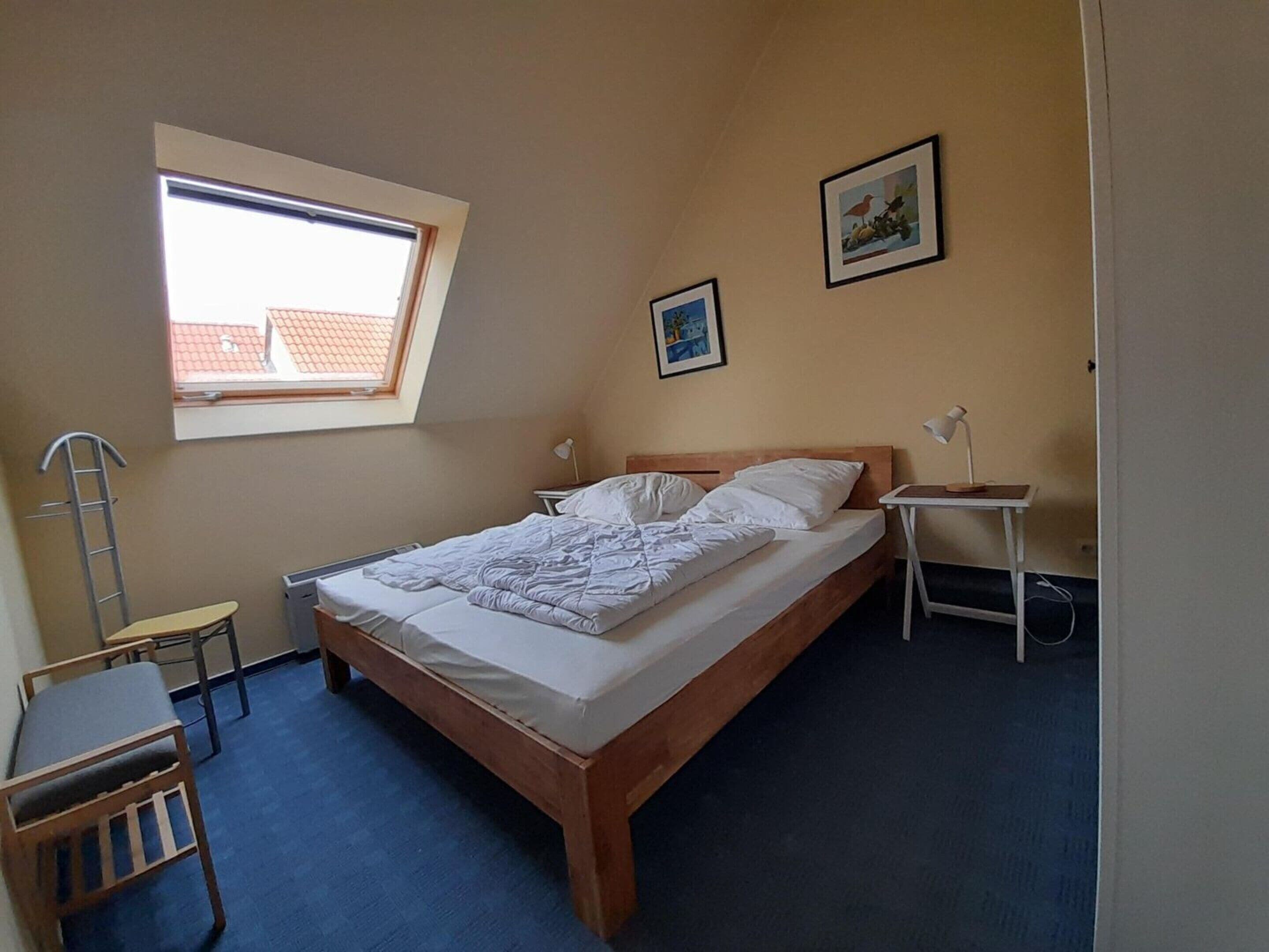 Ein Schlafzimmer mit einem Einzelbett, einem Schreibtisch, einem Stuhl, einem kleinen Tisch mit Lampe, einem Fenster und zwei gerahmten Bildern an der Wand.