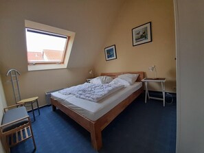 Ein Schlafzimmer mit einem Einzelbett, einem Schreibtisch, einem Stuhl, einem kleinen Tisch mit Lampe, einem Fenster und zwei gerahmten Bildern an der Wand.