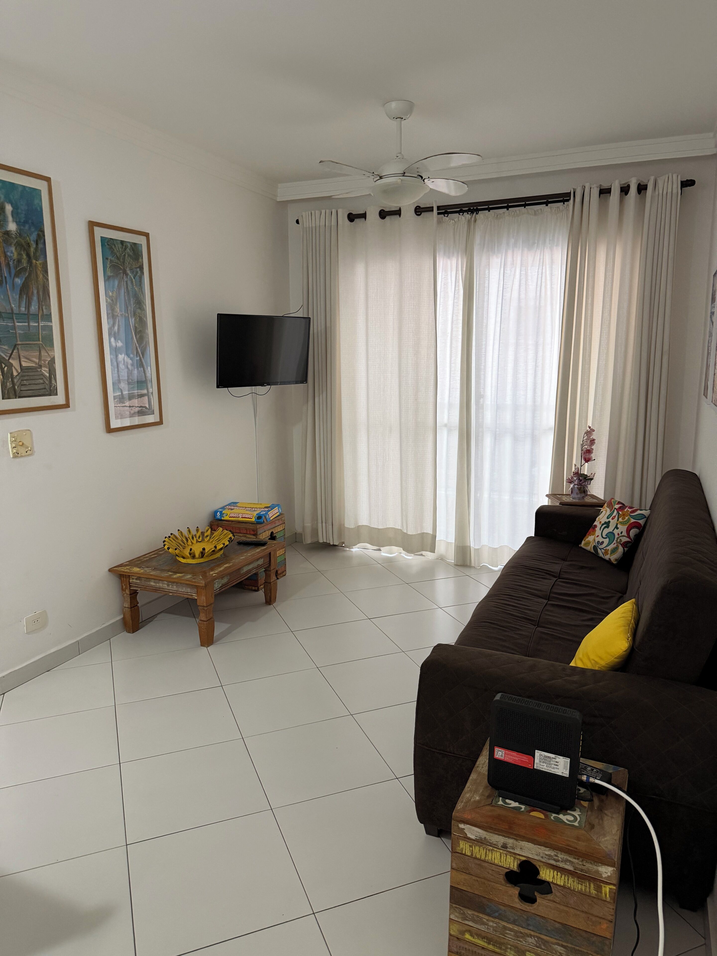 Ein Wohnzimmer mit braunem Sofa, einem an der Wand montierten Fernseher, einem Deckenventilator und einem Fenster mit Vorhängen.