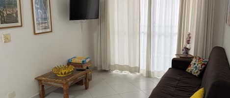 A living room with a brown sofa, a television mounted on the wall, a ceiling fan, and a window with curtains.