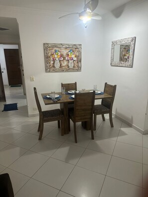 A dining area with a table set for four, chairs, a ceiling fan, and a wall-mounted mirror. - Cozy apartment in Guarujá, Enseada beach. (Guarujá)