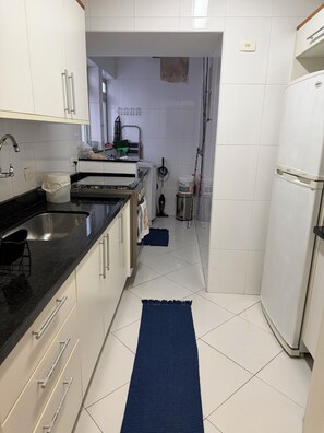 A kitchen with white cabinets, a black countertop, a stainless steel sink, and a blue rug on the floor. - Cozy apartment in Guarujá, Enseada beach. (Guarujá)