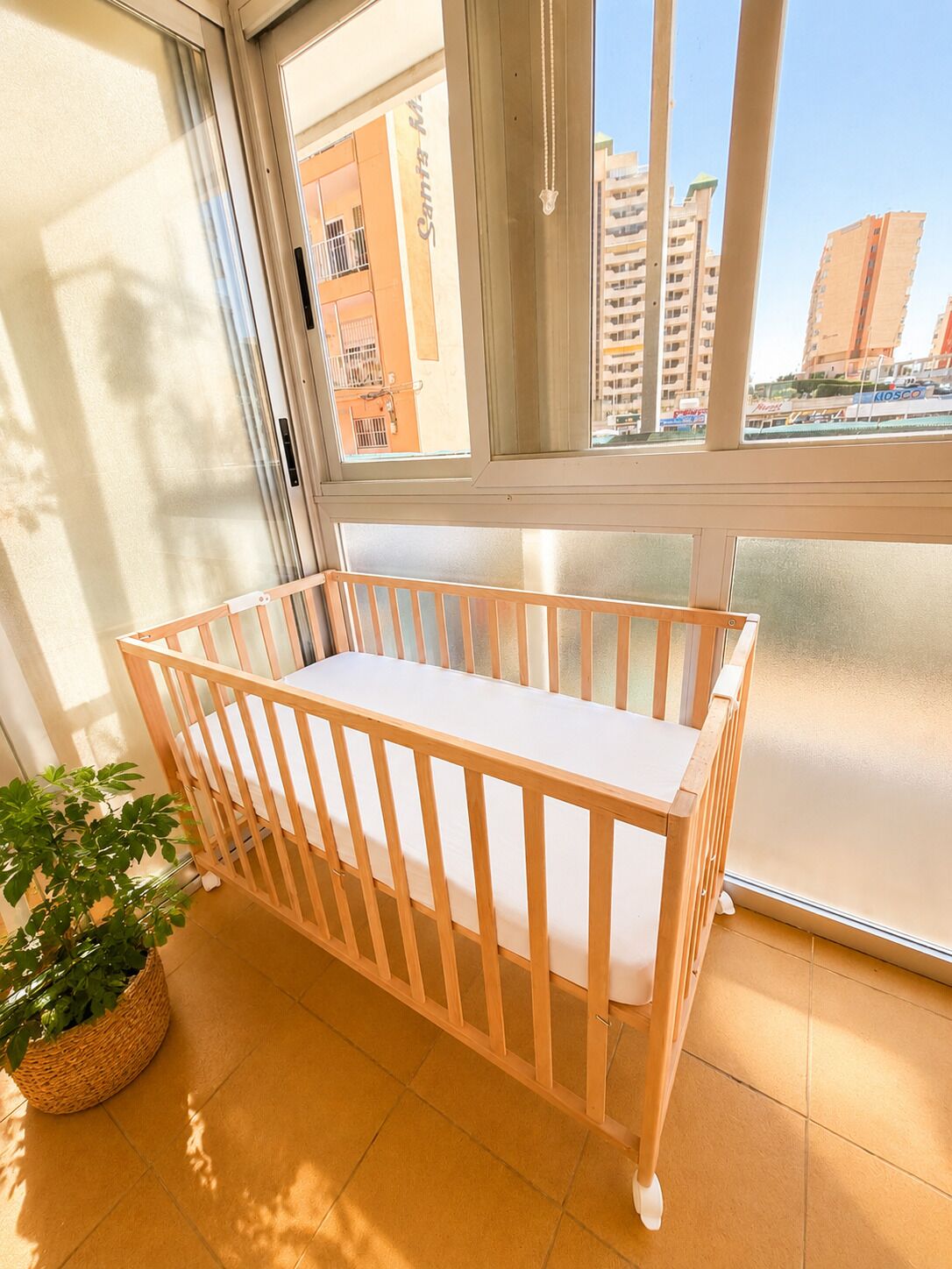 Een kamer met een houten kinderbedje, een potplant en een uitzicht op een stadslandschap door grote ramen.