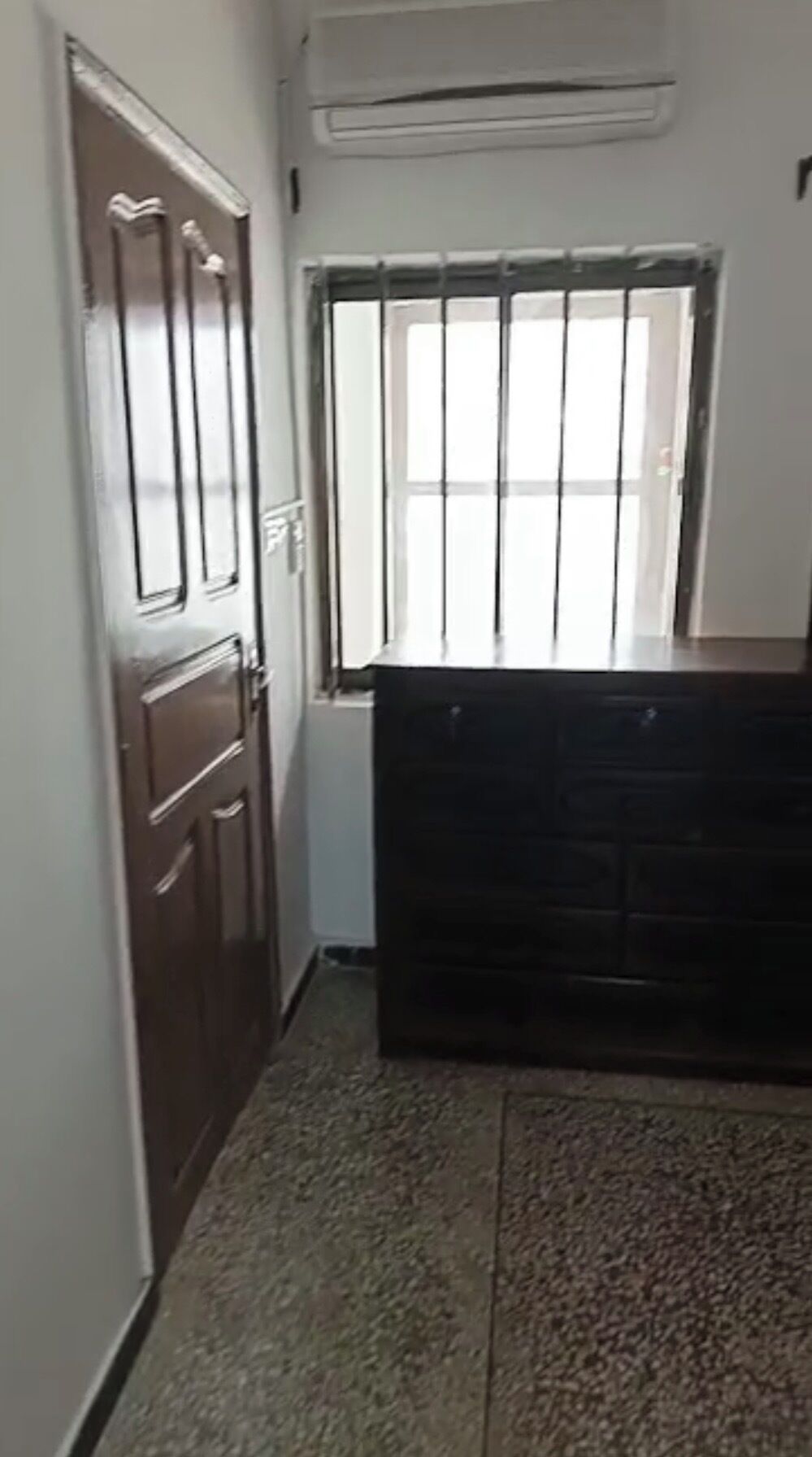Un couloir étroit avec une porte en bois, une fenêtre grillagée et une commode sombre en bois.