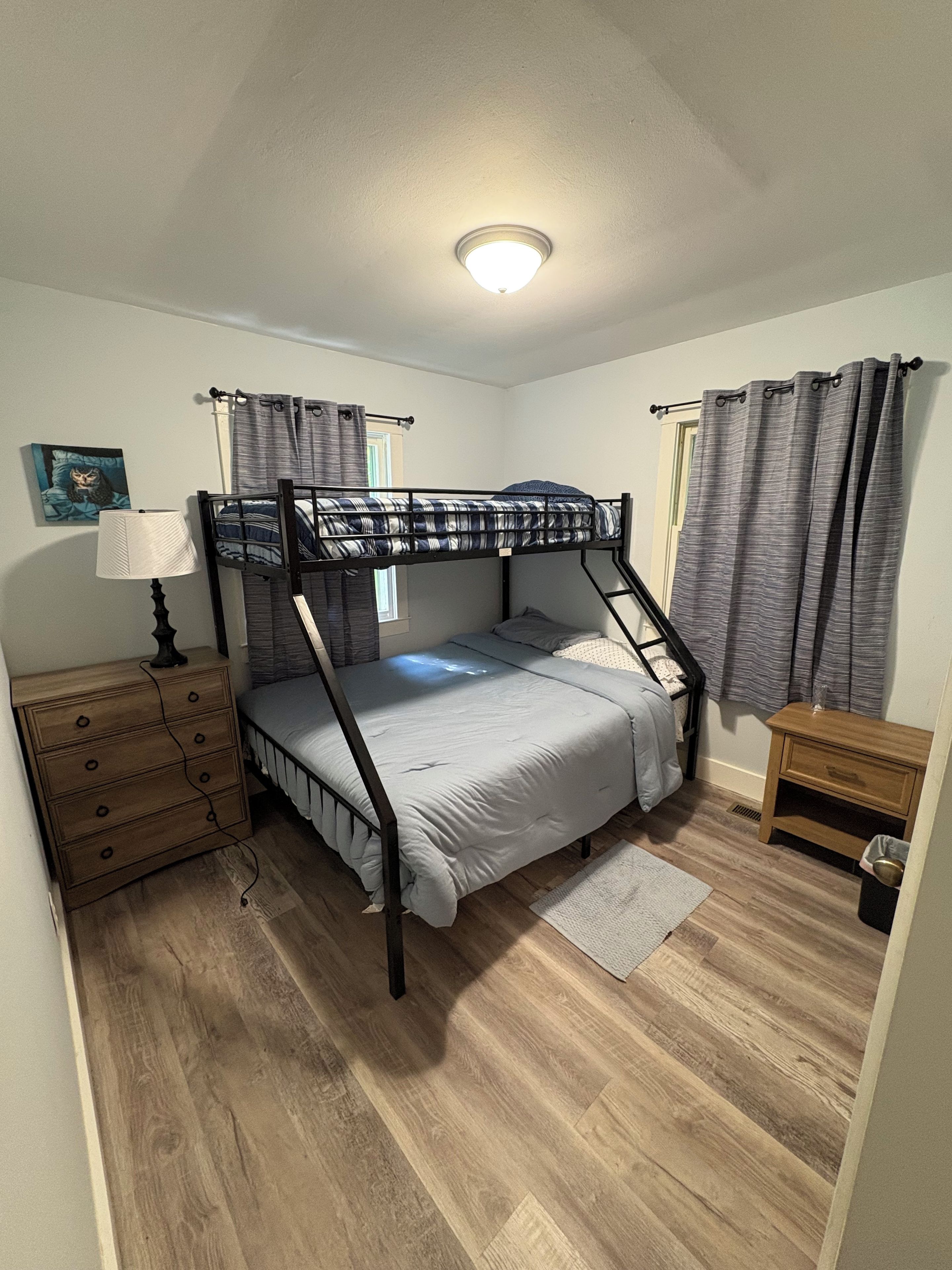 Un lit superposé avec une tête de lit, une table de chevet avec une lampe, une commode en bois, une fenêtre avec des rideaux et un petit tabouret en bois.
