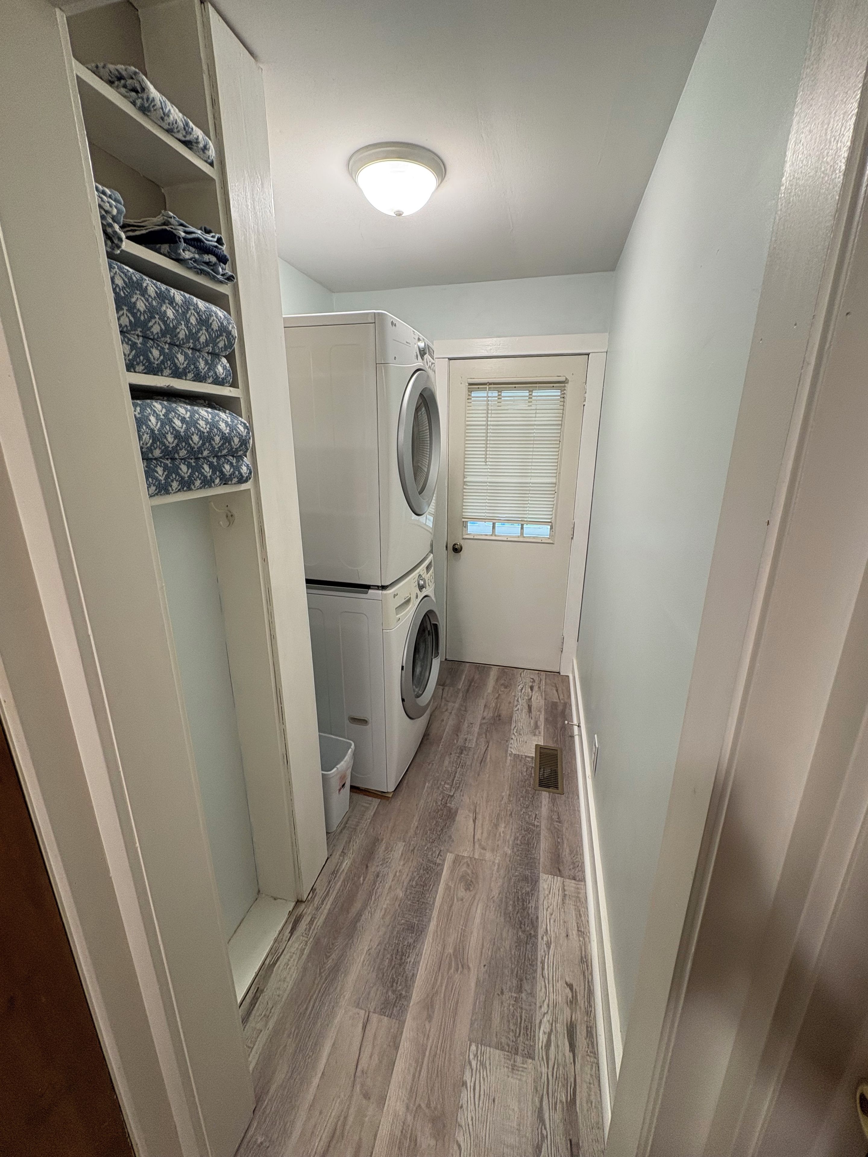 Une buanderie équipée d’un lave-linge et d’un sèche-linge, une petite fenêtre avec des stores et un plancher en bois.