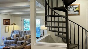 A living room with a brown sofa, a plaid armchair, and a staircase with a black railing.