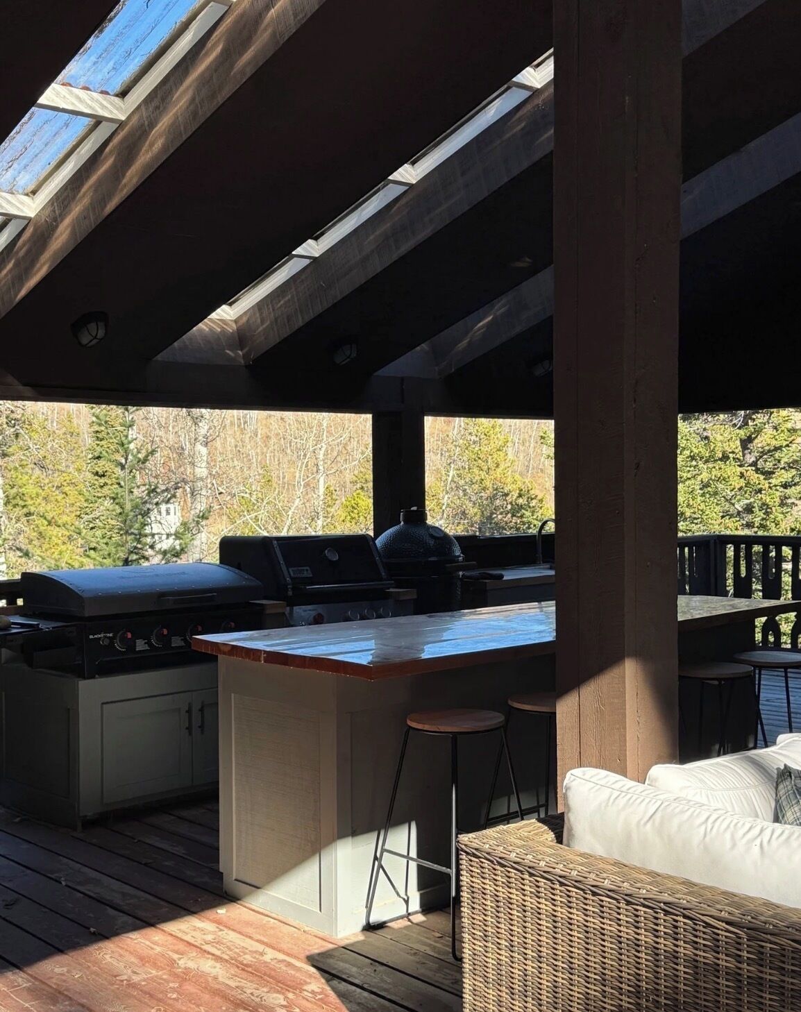 Cocina exterior con parrilla, taburetes de bar y una zona de estar con una silla de mimbre.