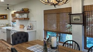 A dining area with a wooden table, chairs, a chandelier, and a view through a window with blinds.
