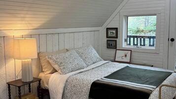 Une chambre avec un plafond en pente, un lit avec une literie à motifs, une lampe de chevet, une fenêtre donnant sur un beau paysage et une petite table avec un livre.