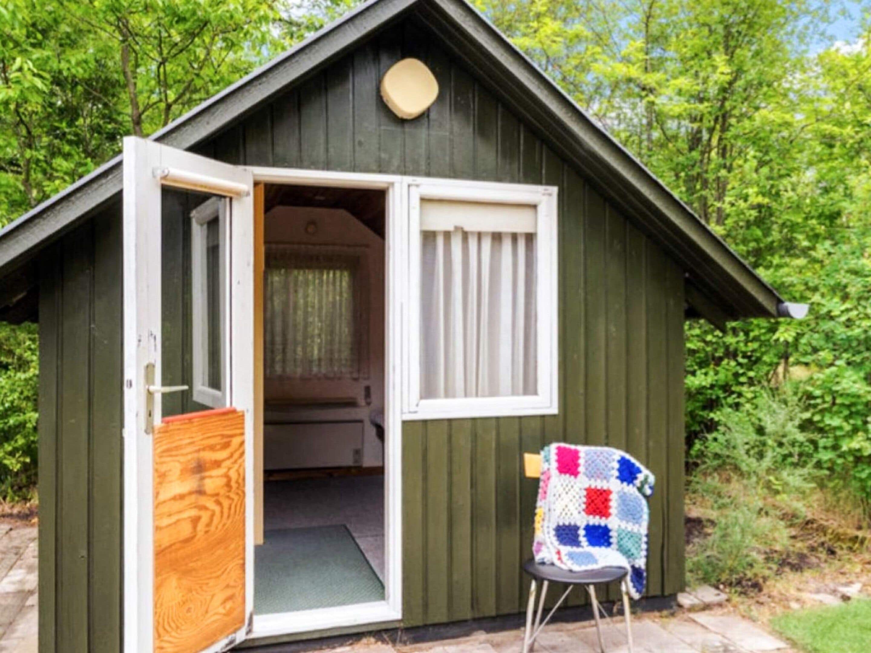 Ein kleines Holzshed mit geöffneter Tür, einer Bank davor und einer Patchworkdecke auf der Bank.