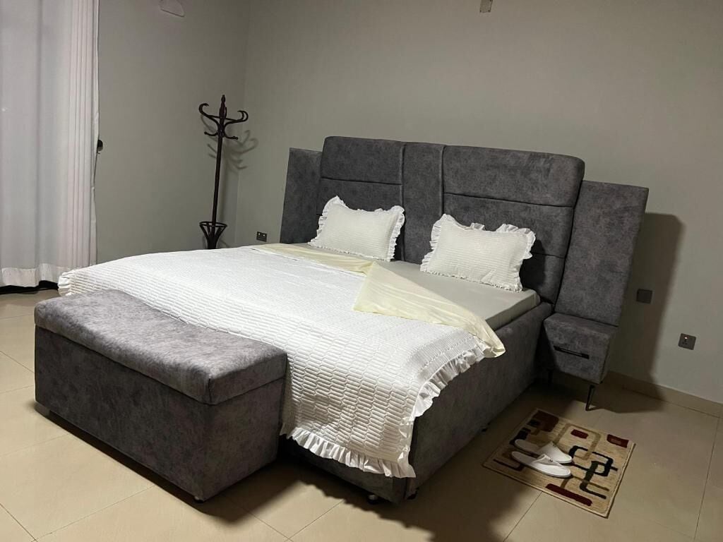 Un dormitorio con cama, un banco tapizado en gris, una mesita de noche y un felpudo.