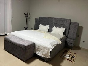 A bedroom with a bed, a grey upholstered bench, a bedside table, and a floor mat. - KAZE CONSOLIDATE HOME INN (Kigali)