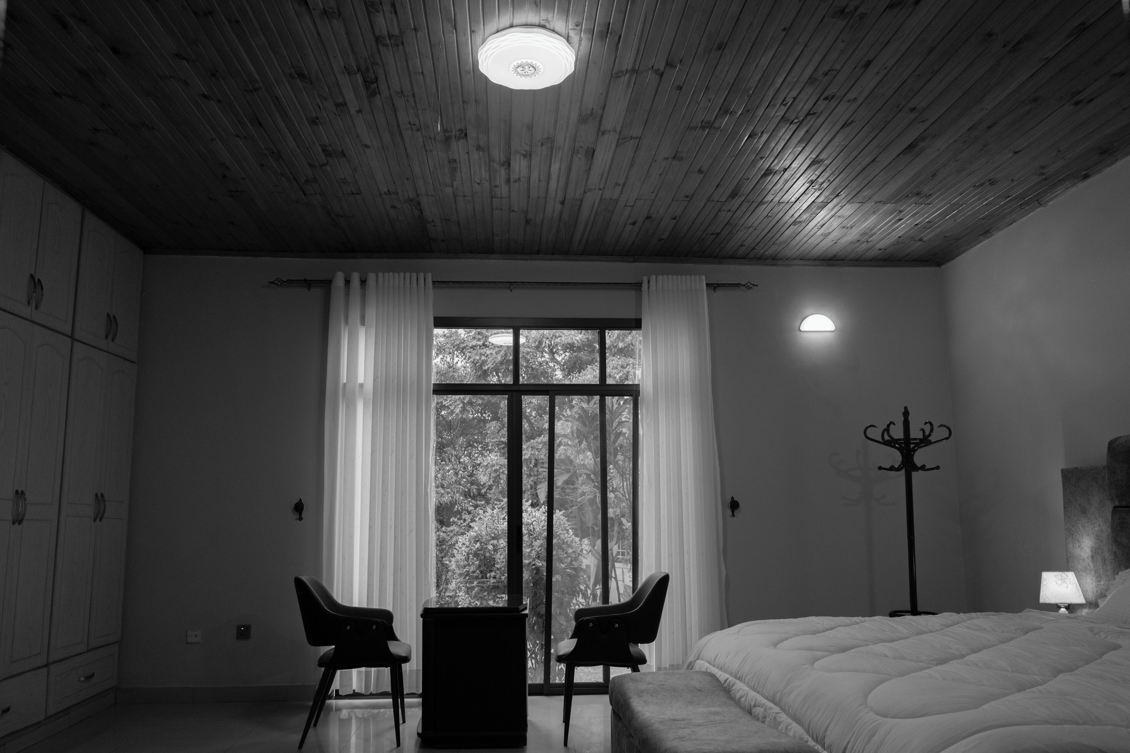 Une pièce avec un plafond en bois, un lit, un bureau avec deux chaises, une fenêtre avec des rideaux et un lampadaire.