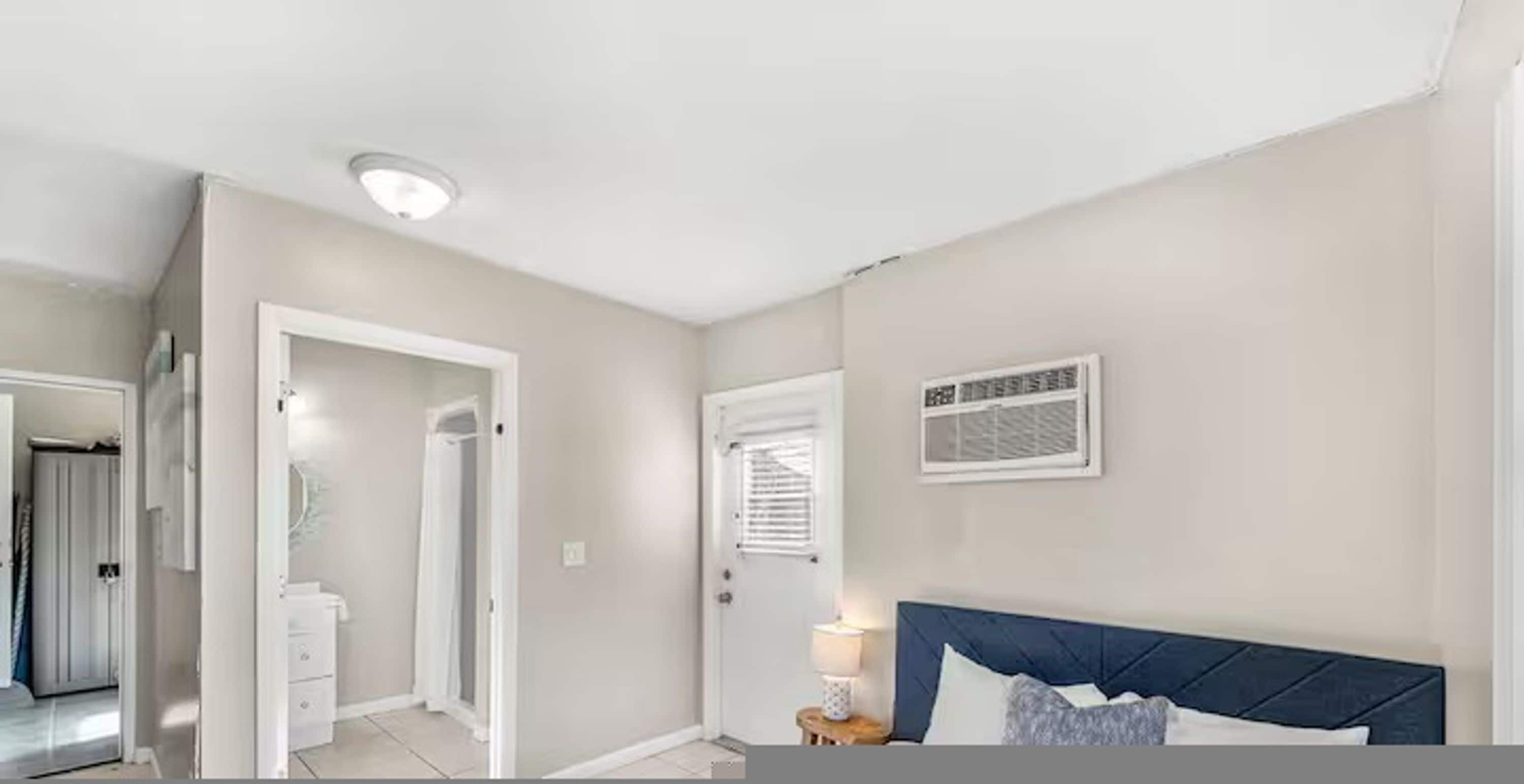 A bedroom with a bed, a nightstand, a window with blinds, and a door leading to another room.