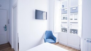 Ein Zimmer mit einem Bett, einem blauen Sessel, einem Wand-Fernseher und einem Fenster mit Blick auf Gebäude.