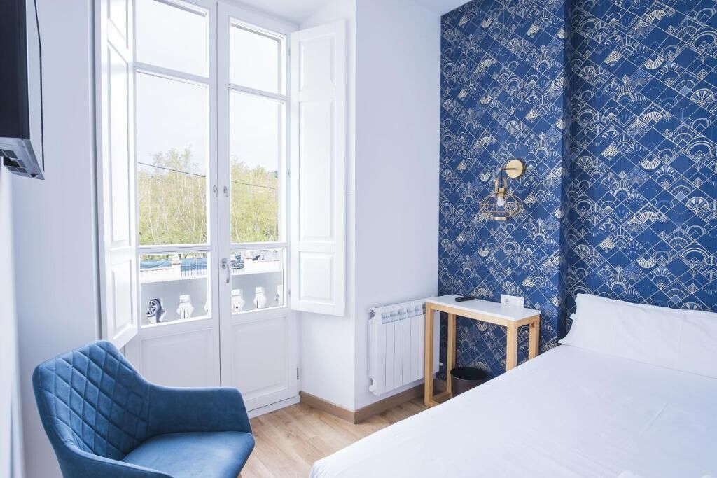 A bedroom with a bed, a blue chair, a small table, a radiator, and a window with a view of greenery.