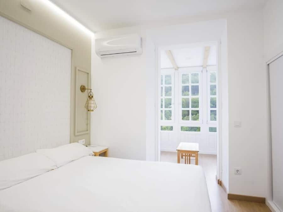 A bedroom with a bed, a bedside table, an air conditioning unit, a window with a view, and a door.