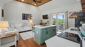 A compact kitchen with a wooden ceiling, a dining area with a white table and chairs, a sofa, and a view of the outdoors.