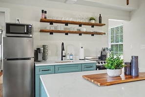 Cocina moderna con refrigerador de acero inoxidable, microondas, estufa y pileta. Hay repisas de madera sobre la mesada con vasos y una planta en maceta.