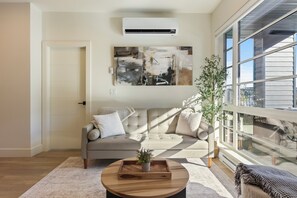 A living room with a sofa, a coffee table with a plant, and a large window. - Bear Mountain Retreat | AC | 400 Sq Ft Patio | Golf & Resort-Style Amenities (Victoria)