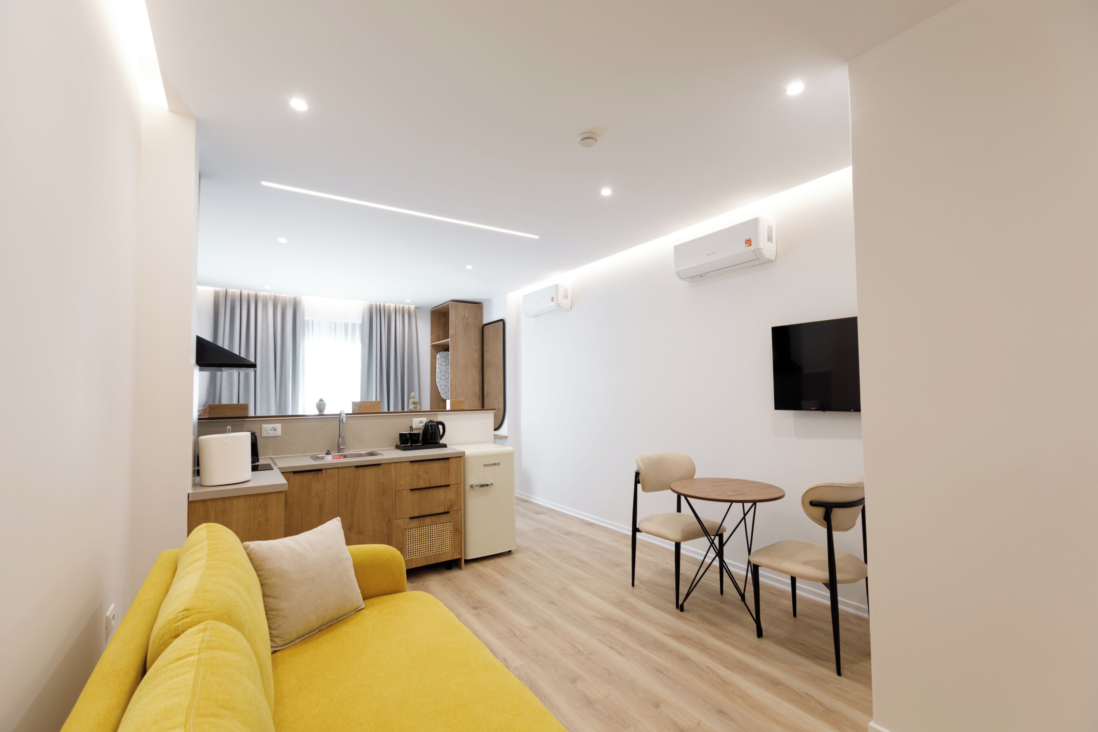 Una sala moderna con un sofá amarillo, un pequeño comedor con dos sillas, una cocina con refrigerador y un televisor montado.