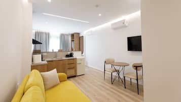 Una sala moderna con un sofá amarillo, un pequeño comedor con dos sillas, una cocina con refrigerador y un televisor montado.