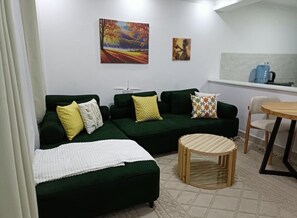 Ein Wohnzimmer mit einem grünen Sofa, einem runden Holztisch und einer dahinterliegenden Küchenzeile.