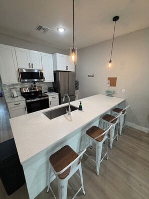 A modern kitchen with white cabinetry, stainless steel appliances, a central island with bar stools, and pendant lighting. - Gorgeous Coastal Townhome - 8 min from Boca Grande, Close to Beaches & Shops (Port Charlotte)