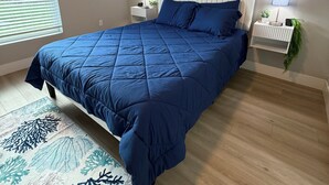 Un dormitorio con cama, dos mesitas de noche, una ventana con persianas y un muro con dos cuadros enmarcados.